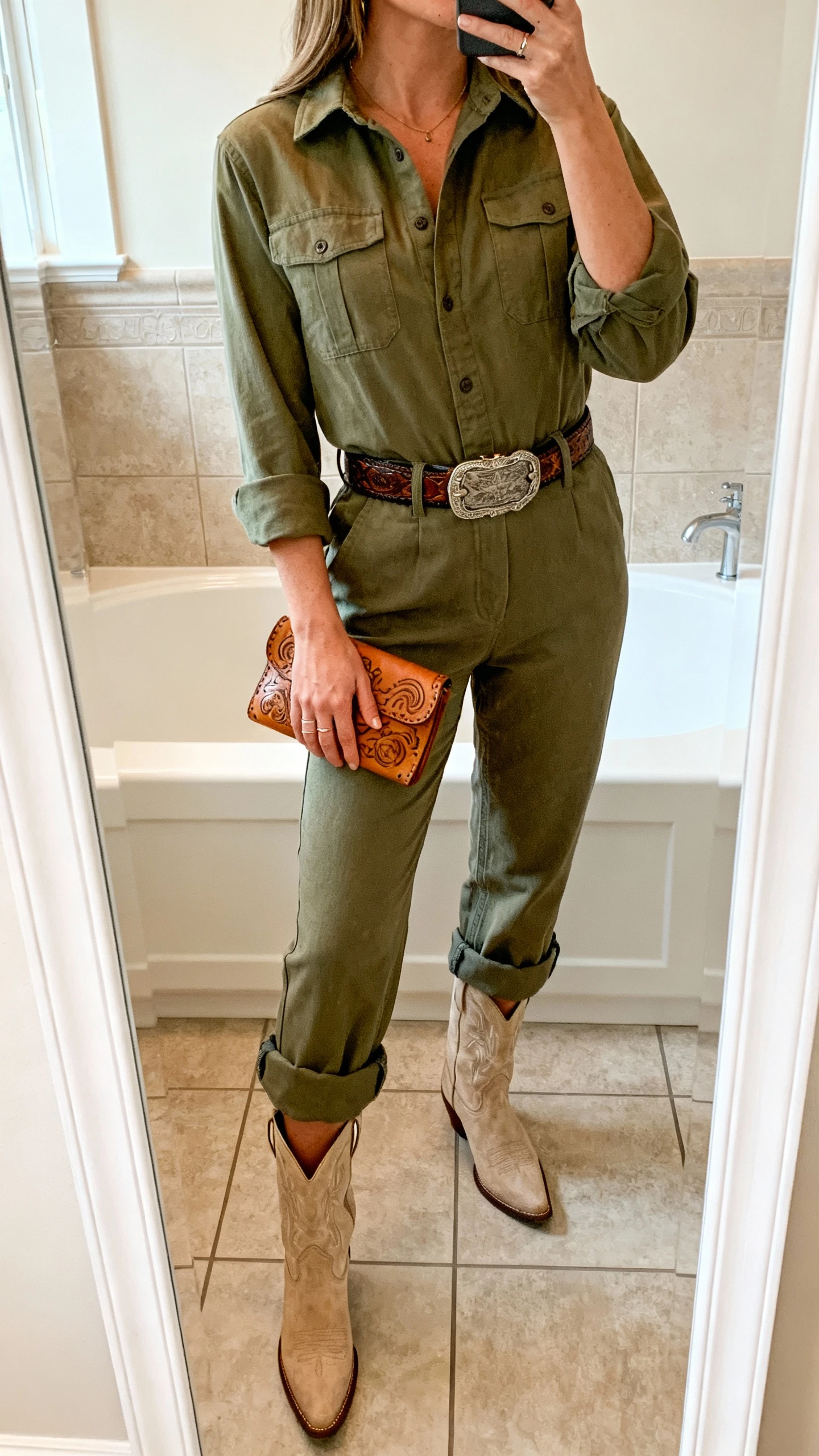 iPhone mirror selfie of a woman in an olive utility jumpsuit cinched with a western belt, rolled cuffs, paired with sand-colored cowboy ankle boots and a tooled leather wristlet; face not visible, cropped at neck, bathroom mirror, indoor lighting, iPhone photo quality.