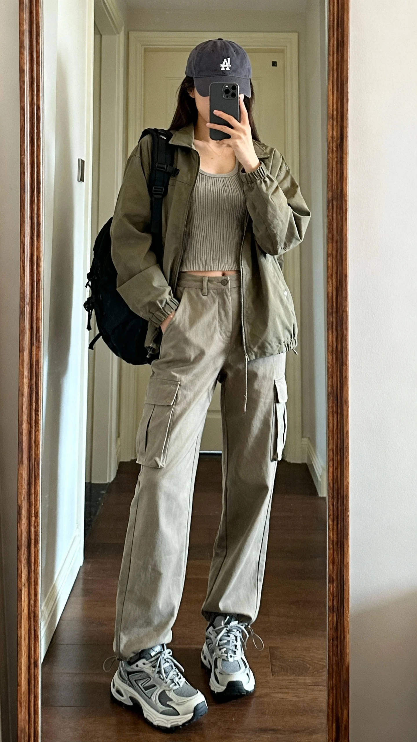 iPhone mirror selfie of a woman in relaxed cargo pants, utility jacket over a ribbed tank, chunky sneakers, baseball cap and backpack, face not visible, entryway mirror, natural daylight, iPhone photo quality.