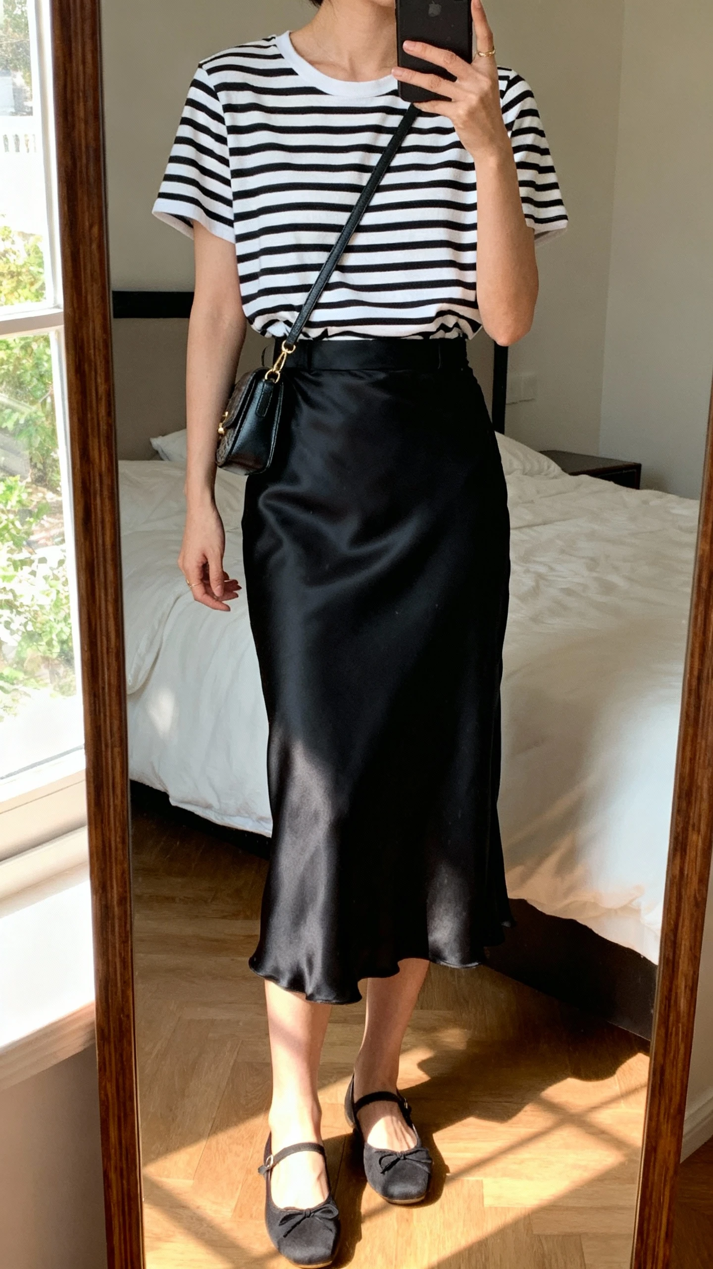 iPhone mirror selfie of a woman wearing a black-and-white Breton striped tee tucked into a black satin slip skirt with simple ballet flats and a small crossbody bag, face not visible, cropped at neck, bedroom mirror, natural window light, iPhone photo quality.