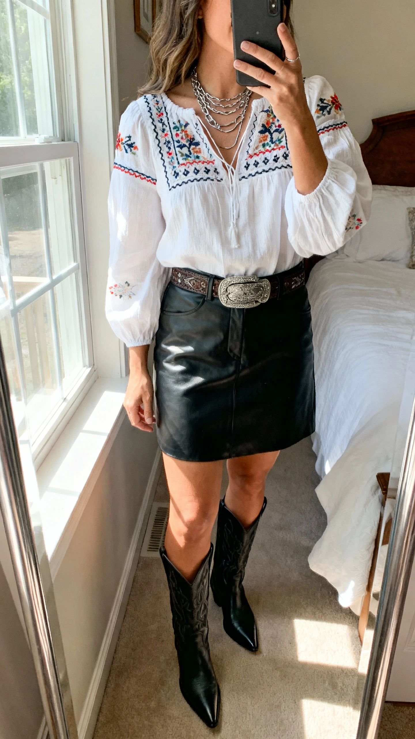 iPhone mirror selfie of a woman wearing a black leather skort with an embroidered white peasant top, western belt, black pointed-toe cowboy boots, and layered silver necklaces; face not visible, side angle, bedroom mirror, natural window light, iPhone photo quality.
