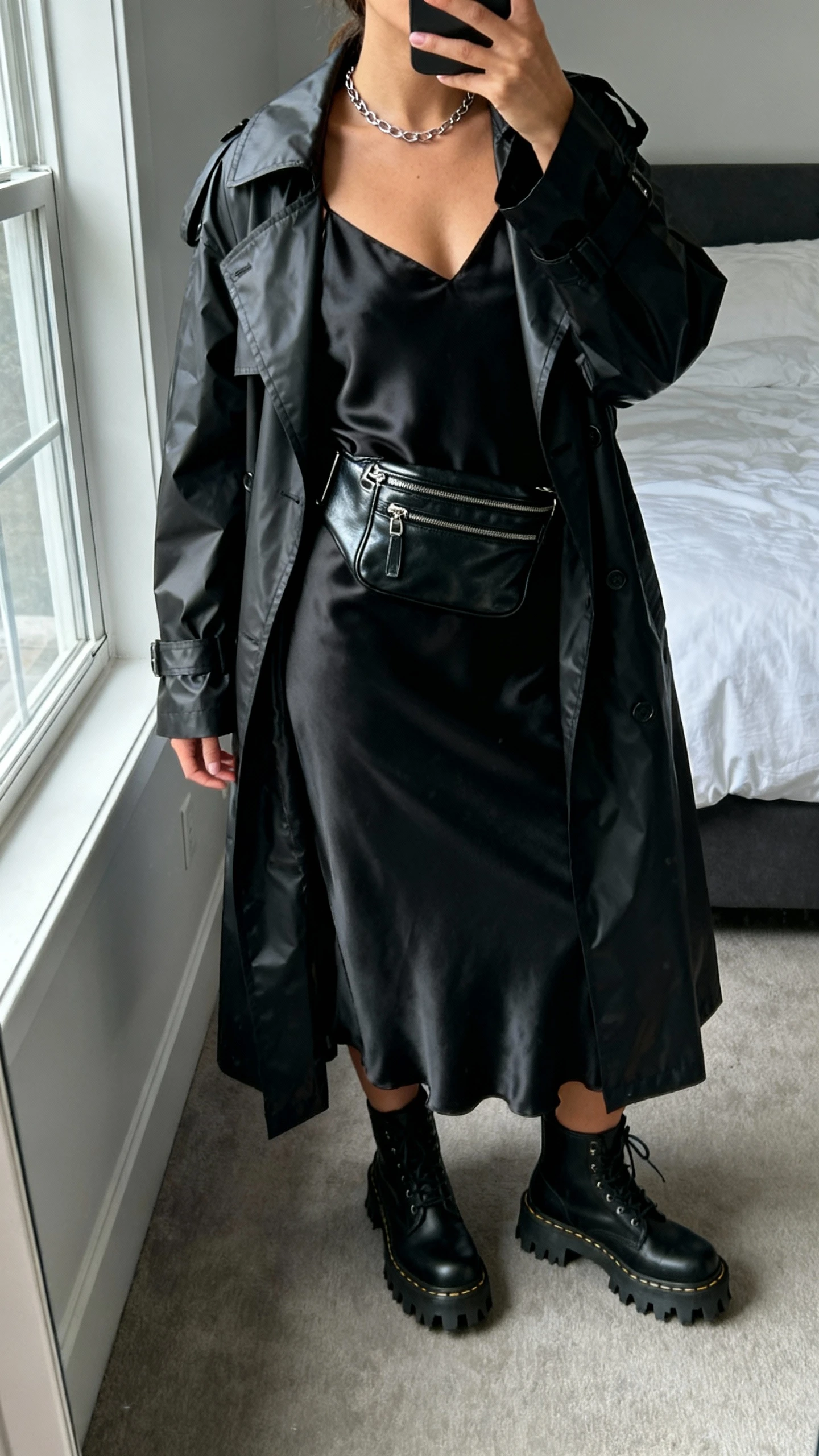 iPhone mirror selfie of a woman wearing a black satin slip dress under a matte black waterproof trench, lug-sole boots, leather moto-style belt bag, minimal chain necklace, face not visible, cropped at shoulders, bedroom mirror, overcast window light, iPhone photo quality.
