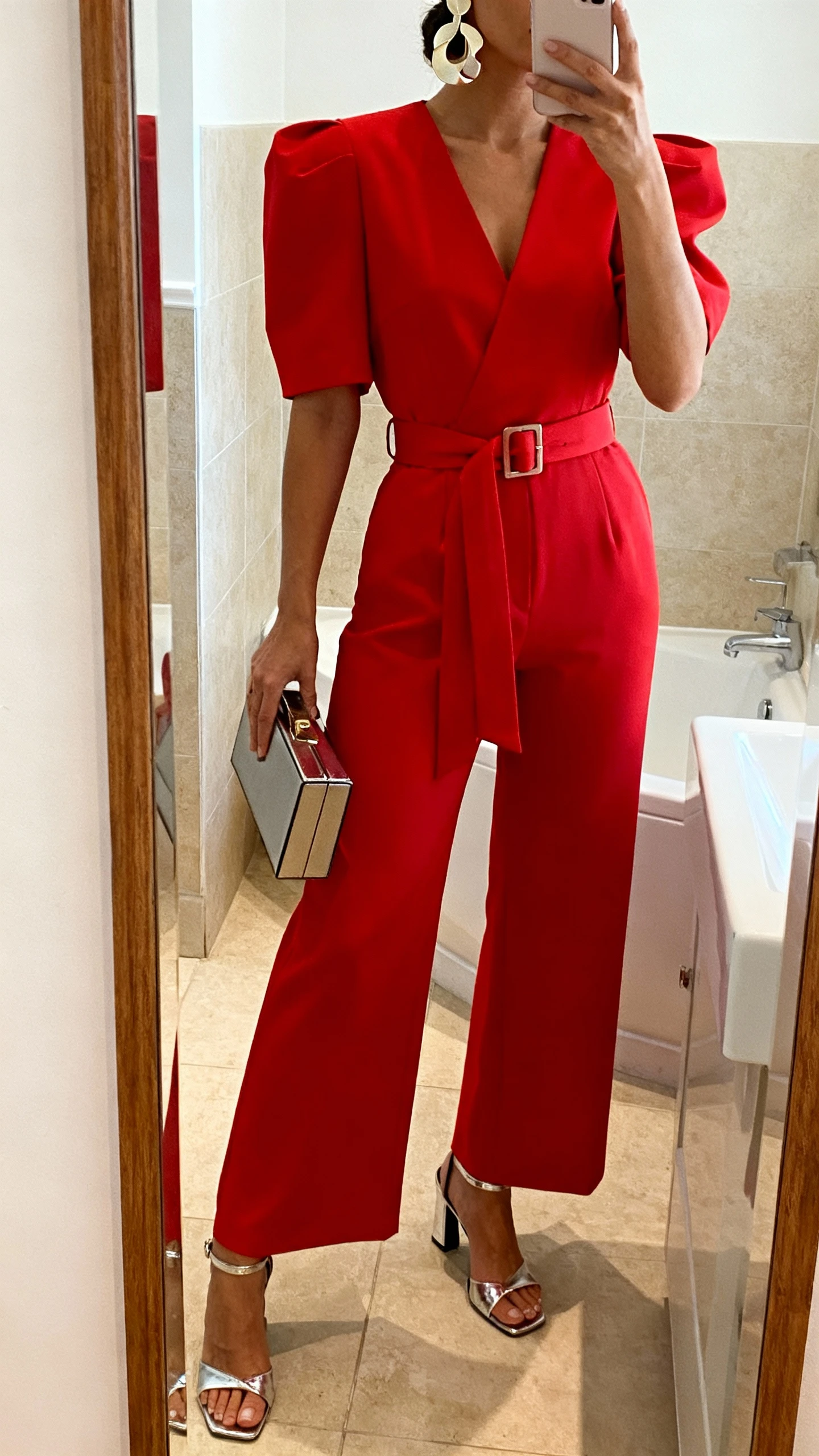 iPhone mirror selfie of a woman wearing a bold red belted statement jumpsuit with strong shoulders and wide legs, metallic heeled sandals, box clutch, sculptural earrings, face not visible, bathroom mirror, warm indoor lighting, iPhone photo quality.