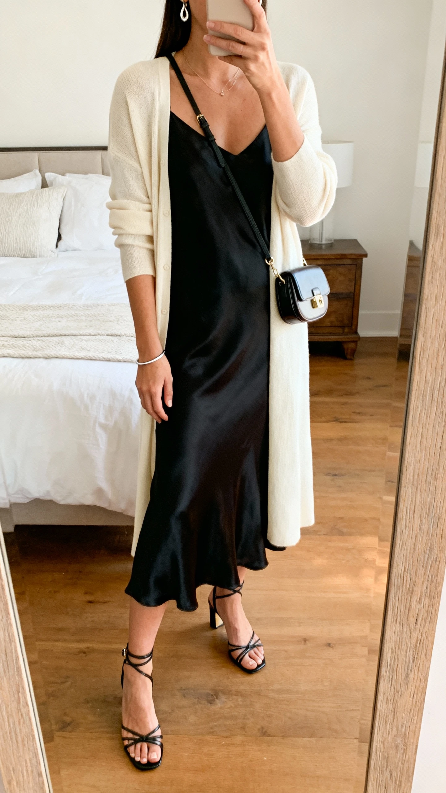 iPhone mirror selfie of a woman wearing a cream mid-length cardigan over a black midi silk slip dress, strappy heeled sandals, a compact shoulder bag, delicate drop earrings, and a thin cuff bracelet, face not visible, cropped at neck, bedroom mirror with warm indoor lighting, iPhone photo quality.