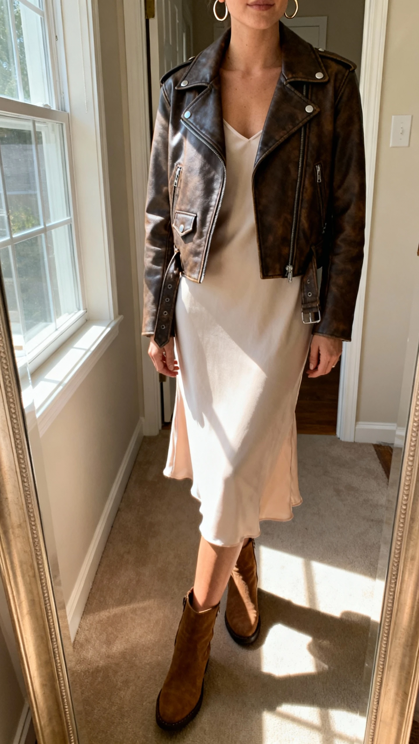 iPhone mirror selfie of a woman wearing a leather biker jacket over a soft slip dress, ankle boots, and subtle hoop earrings, face not visible, cropped at shoulders, entryway mirror, natural window light, iPhone photo quality.