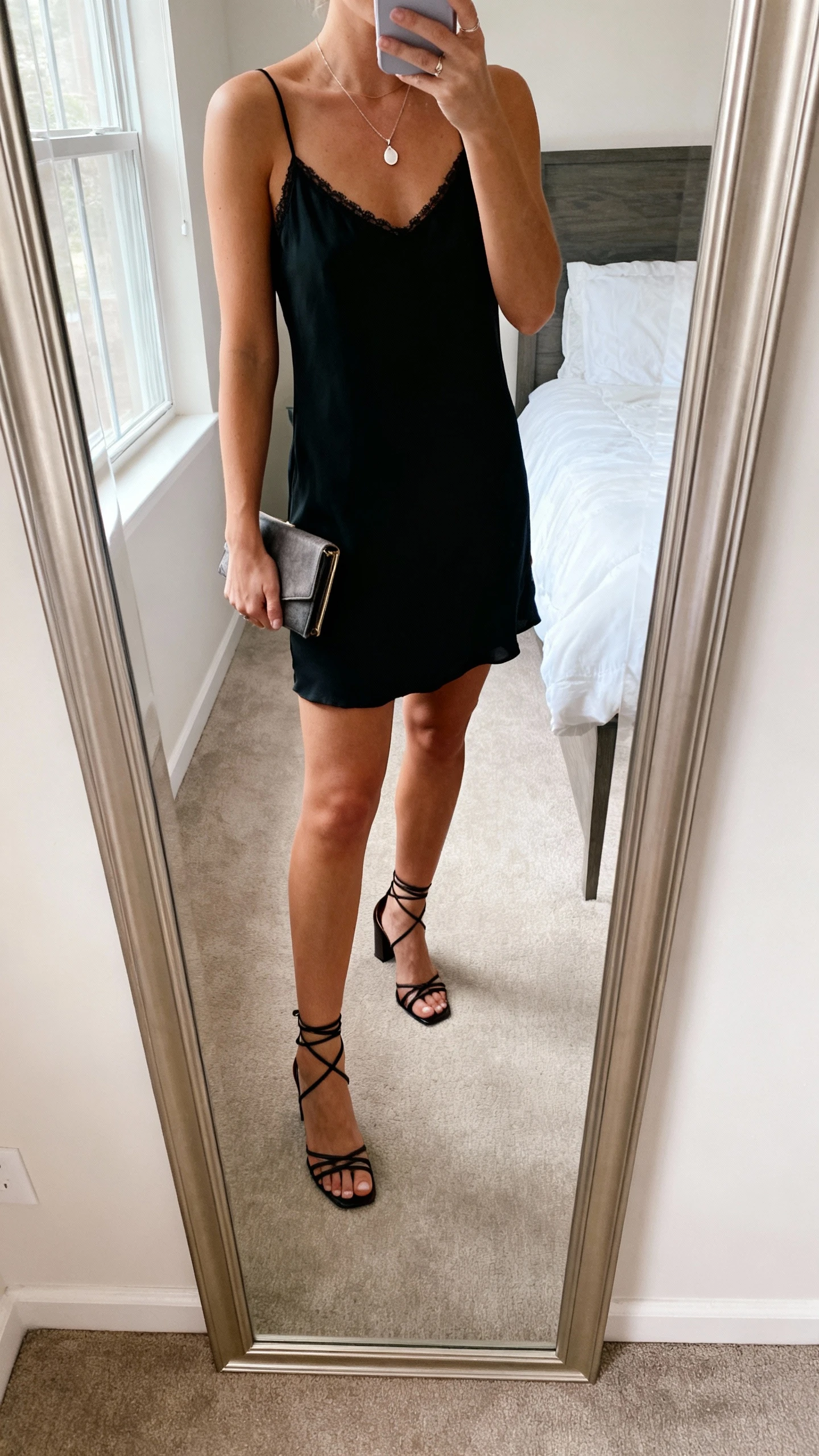 iPhone mirror selfie of a woman wearing a little black slip dress with delicate straps, strappy heeled sandals, a dainty pendant necklace, and a small clutch, face not visible, cropped at neck, bedroom mirror, natural window light, iPhone photo quality.