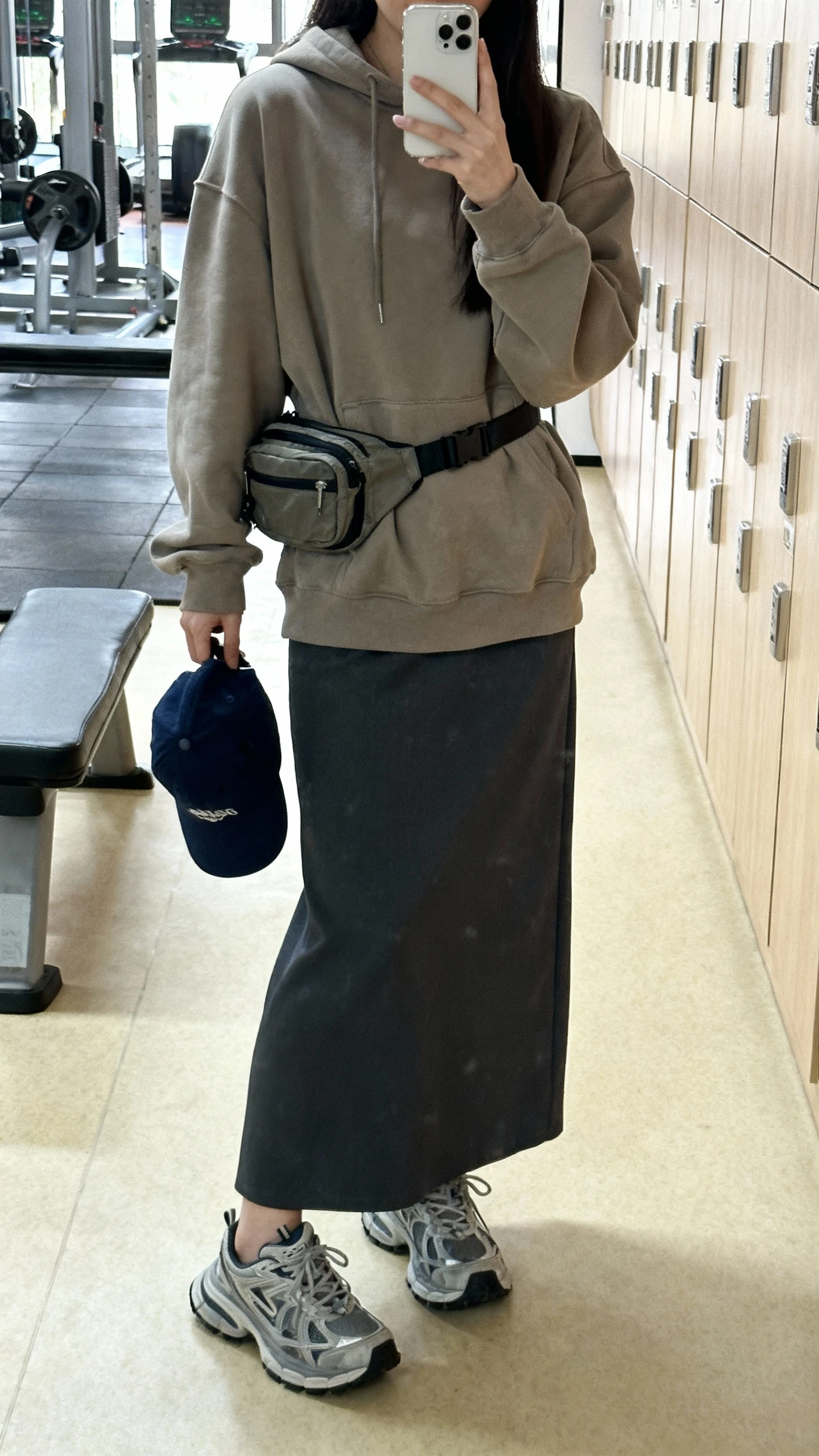 iPhone mirror selfie of a woman wearing a long skirt with a slouchy hoodie and chunky sneakers, baseball cap in hand and a belt bag, face not visible, cropped at shoulders, gym locker mirror, bright indoor lighting, iPhone photo quality.