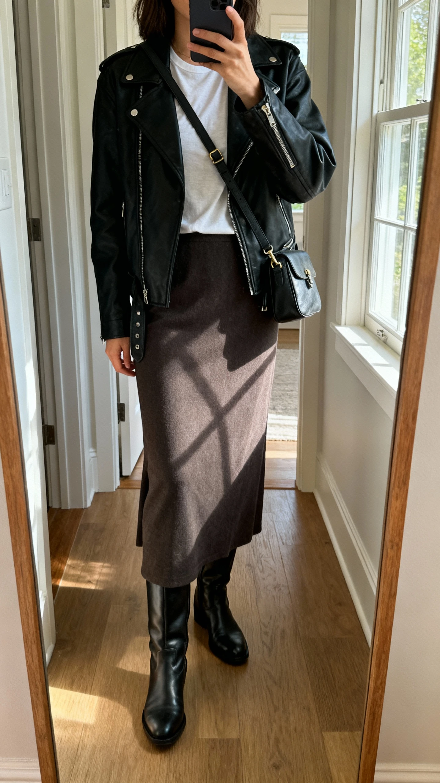 iPhone mirror selfie of a woman wearing a long skirt with knee-high boots and a black moto jacket over a simple tee, crossbody bag, face not visible, cropped at shoulders, entryway full-length mirror, morning window light, iPhone photo quality.