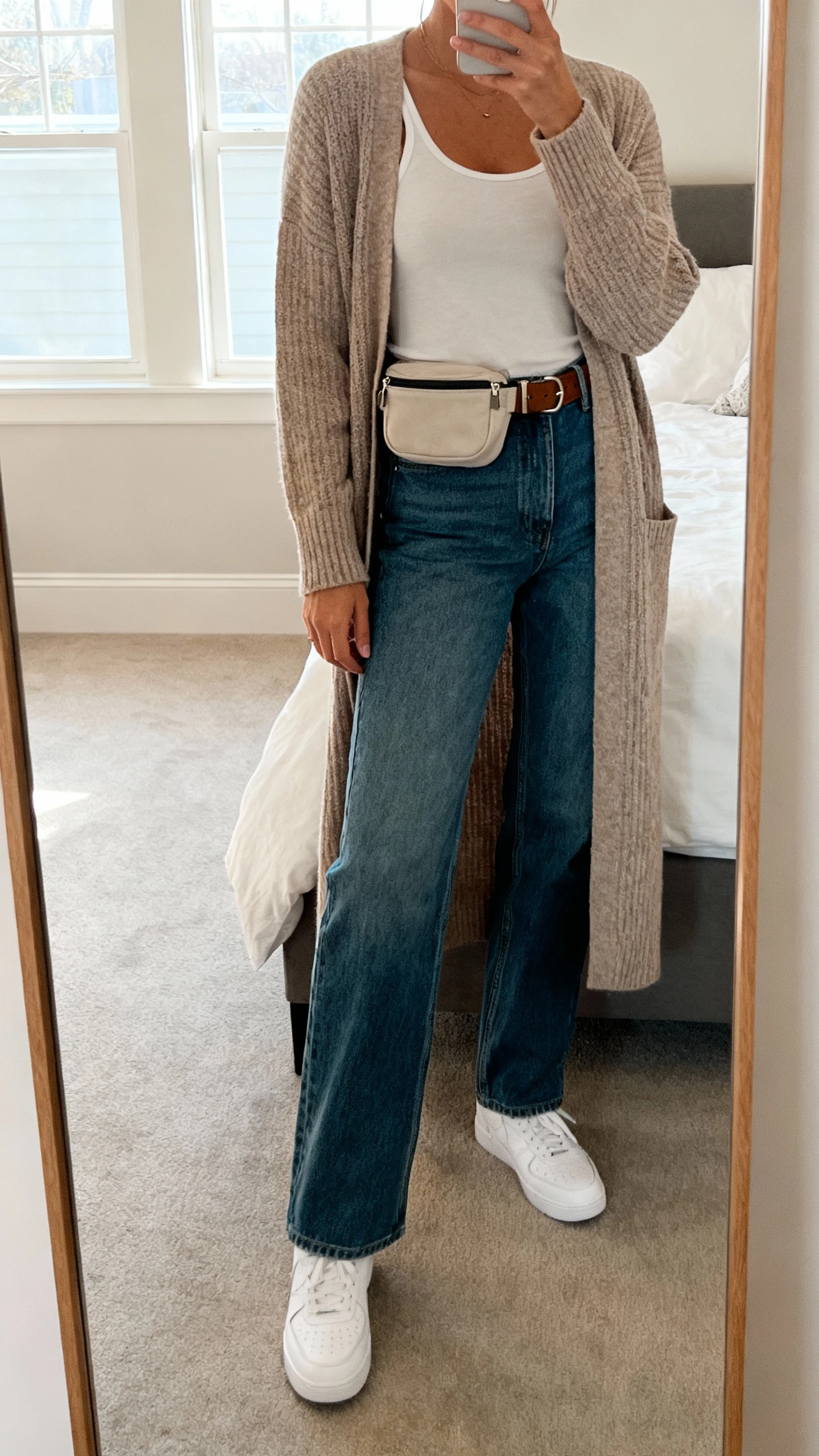 iPhone mirror selfie of a woman wearing a longline cardigan, straight-leg jeans, simple tank, white sneakers, belt bag, face not visible, cropped at shoulders, bedroom mirror with soft window light, iPhone photo quality.