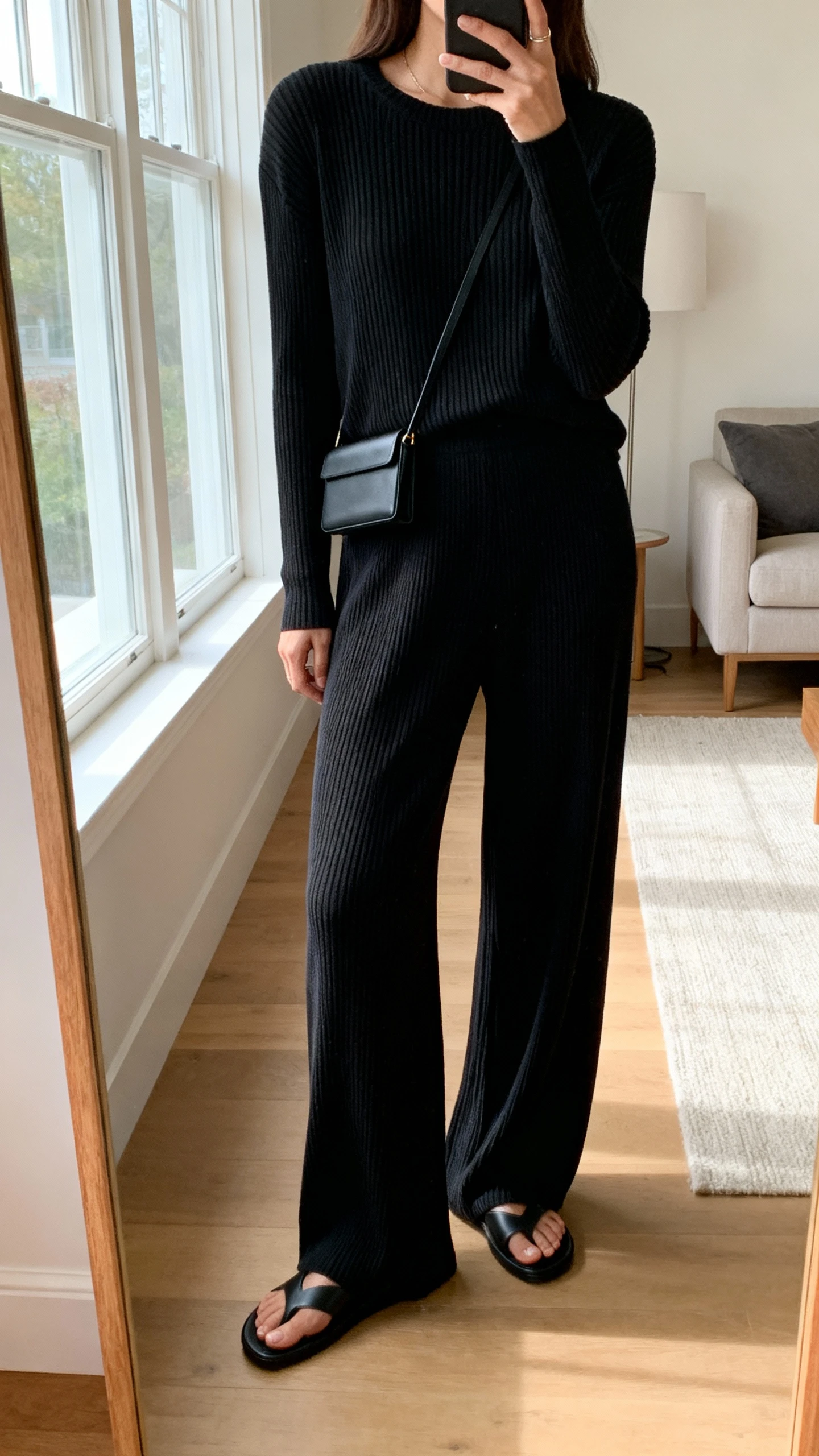 iPhone mirror selfie of a woman wearing a luxe black ribbed knit top with matching flowy black knit pants, black slide sandals, and a slim black crossbody, face not visible, cropped at shoulders, living room mirror, morning window light, iPhone photo quality.