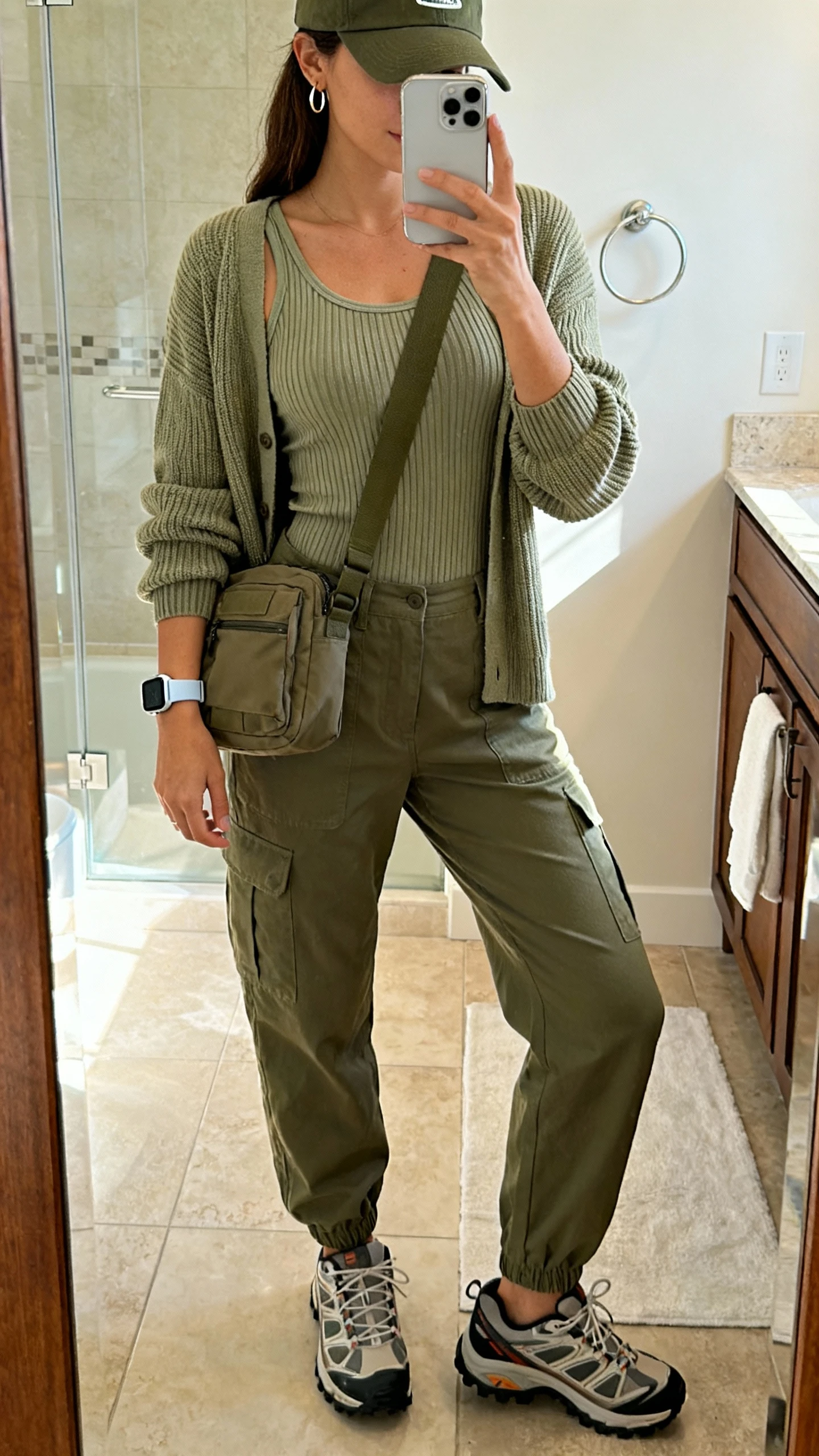 iPhone mirror selfie of a woman wearing a matching ribbed knit set (fitted tank and coordinating cardigan), relaxed-fit olive cargo pants, retro trail sneakers, a utility crossbody, a baseball cap, minimal hoops, and a sporty watch, face not visible, cropped at shoulders, bathroom mirror with soft morning light, iPhone photo quality.