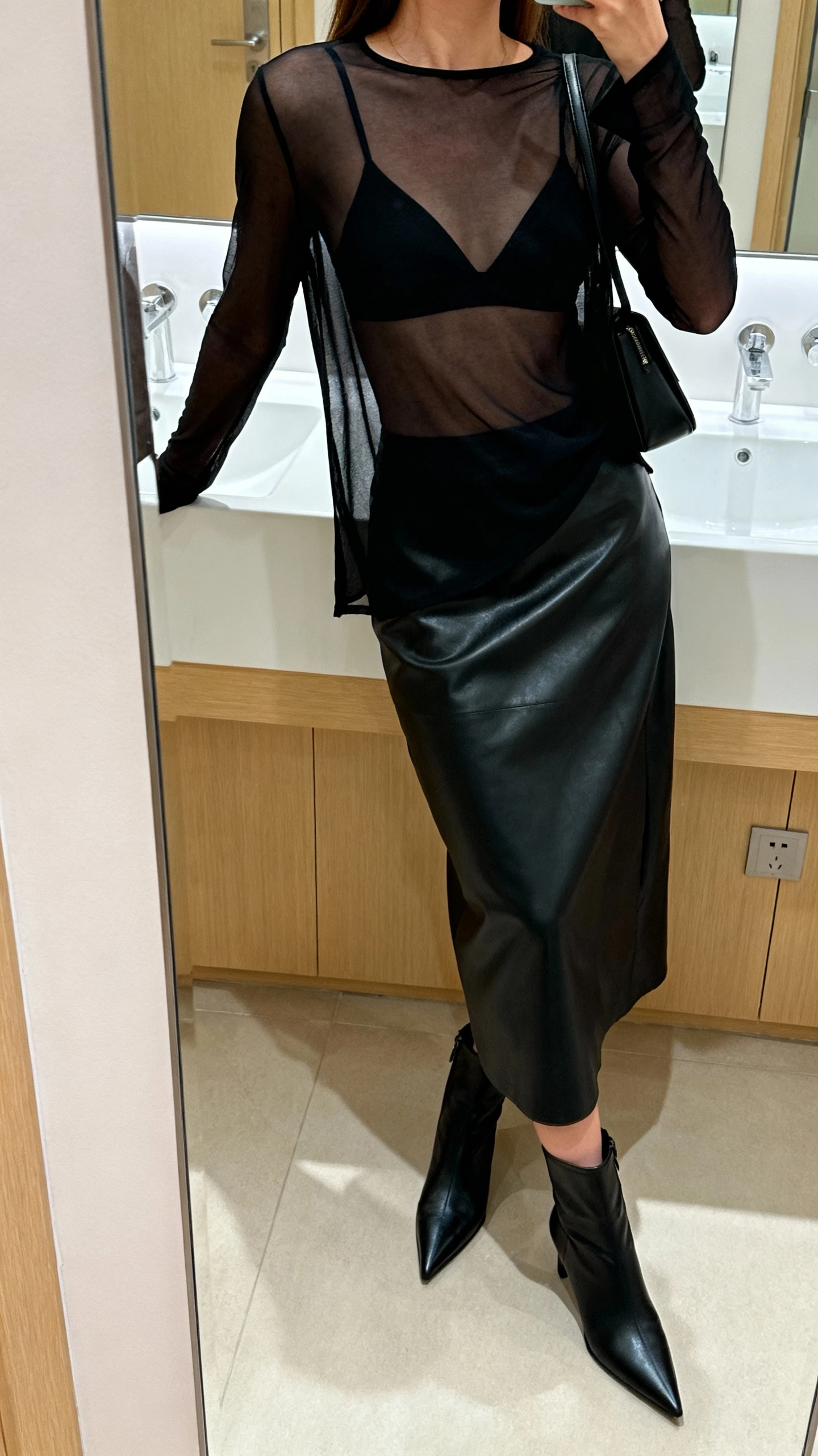 iPhone mirror selfie of a woman wearing a sheer black long-sleeve top over a black bralette, black leather midi skirt, pointed black ankle boots, and minimal black shoulder bag, face not visible, side angle, bathroom mirror, soft indoor lighting, iPhone photo quality.