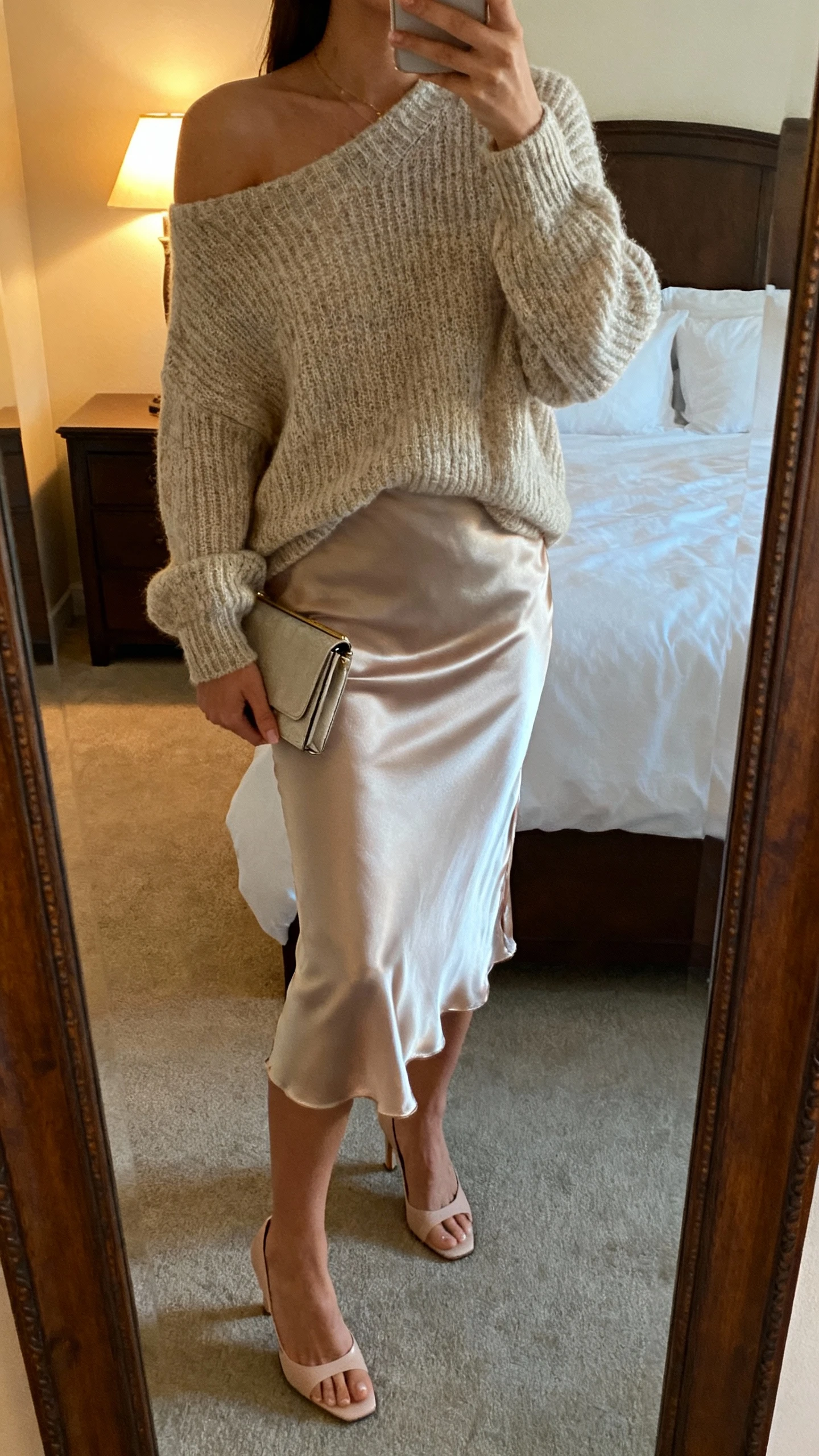 iPhone mirror selfie of a woman wearing a silky slip skirt with an off-shoulder knit sweater, delicate heels, and a small clutch, face not visible, cropped at neck, bedroom mirror, soft evening lamp light, iPhone photo quality.
