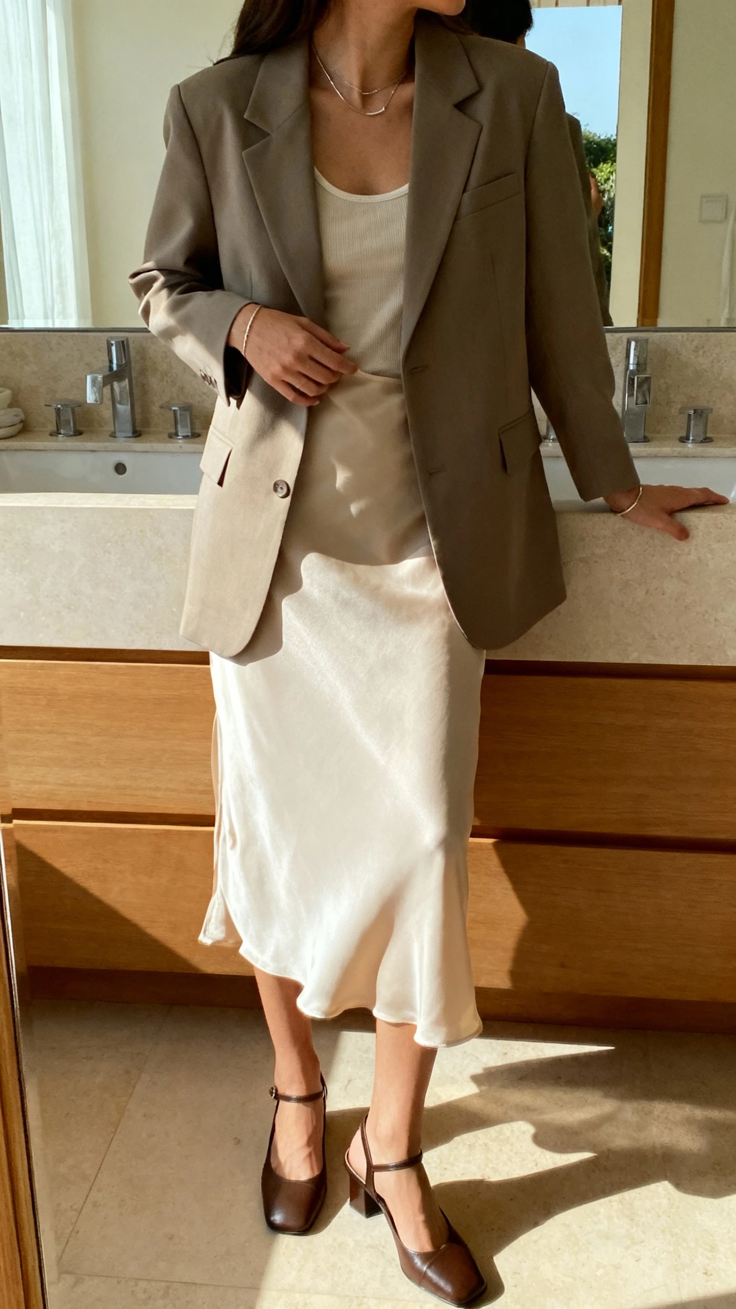 iPhone mirror selfie of a woman wearing a slip skirt with a matching tailored blazer and simple tank, slingback heels, and minimalist jewelry, face not visible, side angle, bathroom mirror, natural morning light, iPhone photo quality.