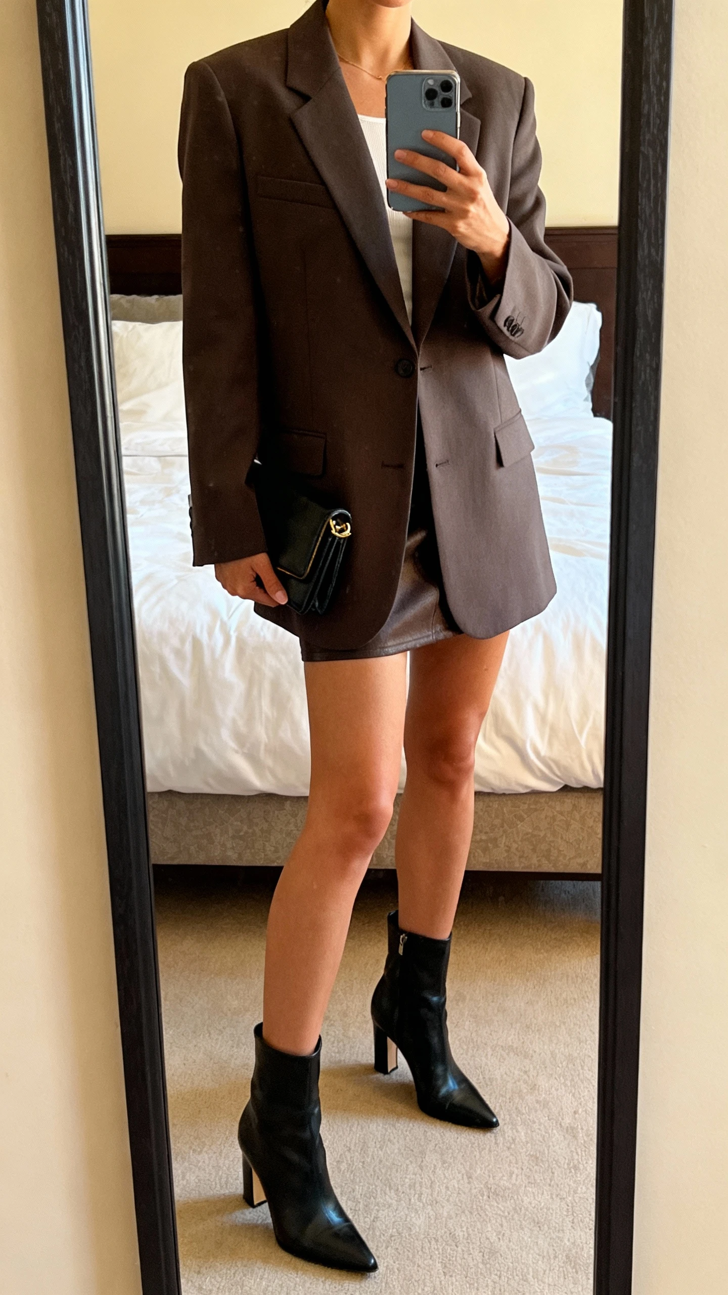 iPhone mirror selfie of a woman wearing a structured power blazer with a mini skirt, heeled ankle boots, and a clutch, face not visible, cropped at neck, bedroom mirror, warm indoor lighting, iPhone photo quality.