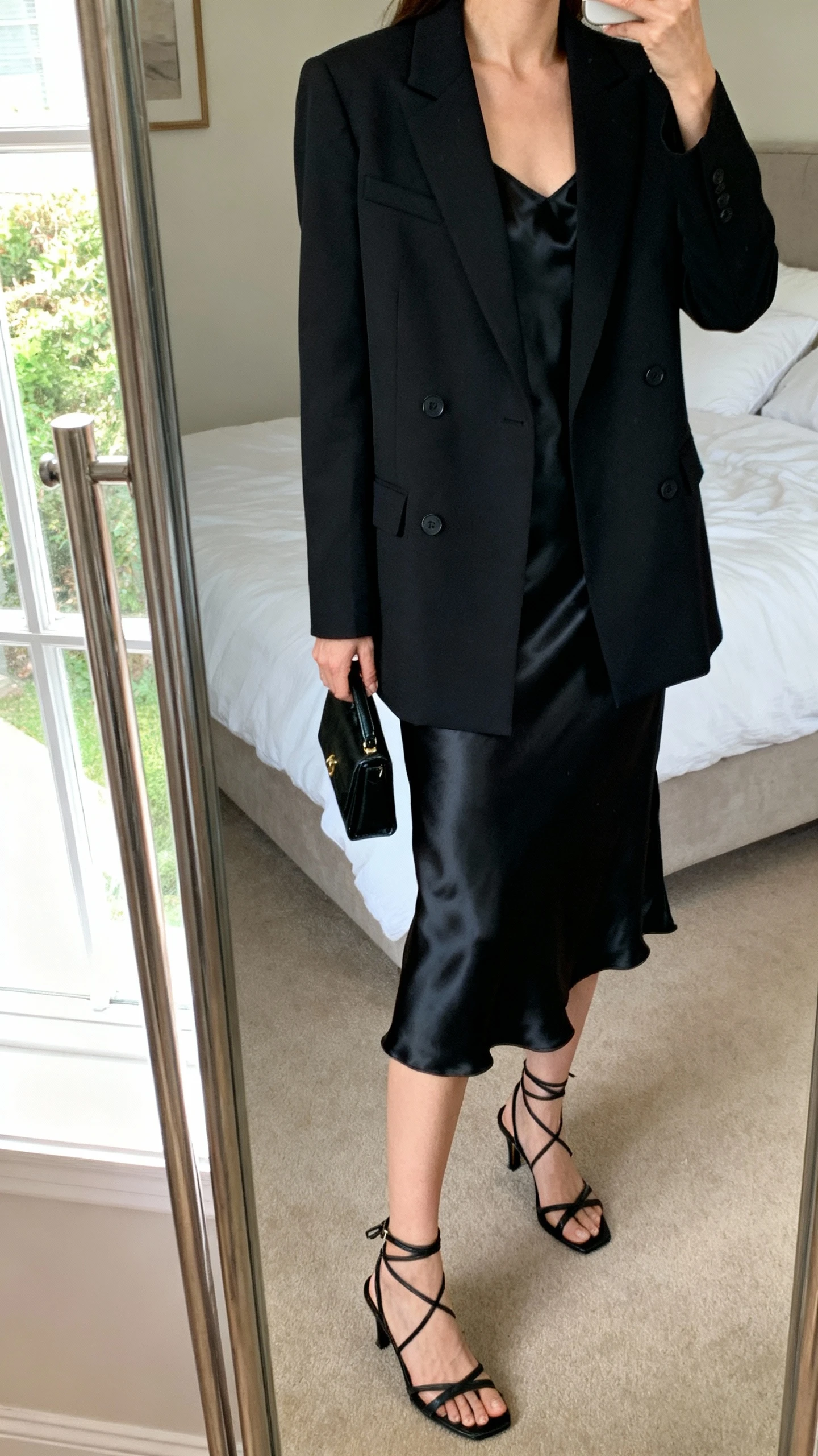 iPhone mirror selfie of a woman wearing a tailored black blazer over a black satin slip dress with strappy black heels and a small black clutch, face not visible, cropped at neck, bedroom mirror, natural window light, iPhone photo quality.