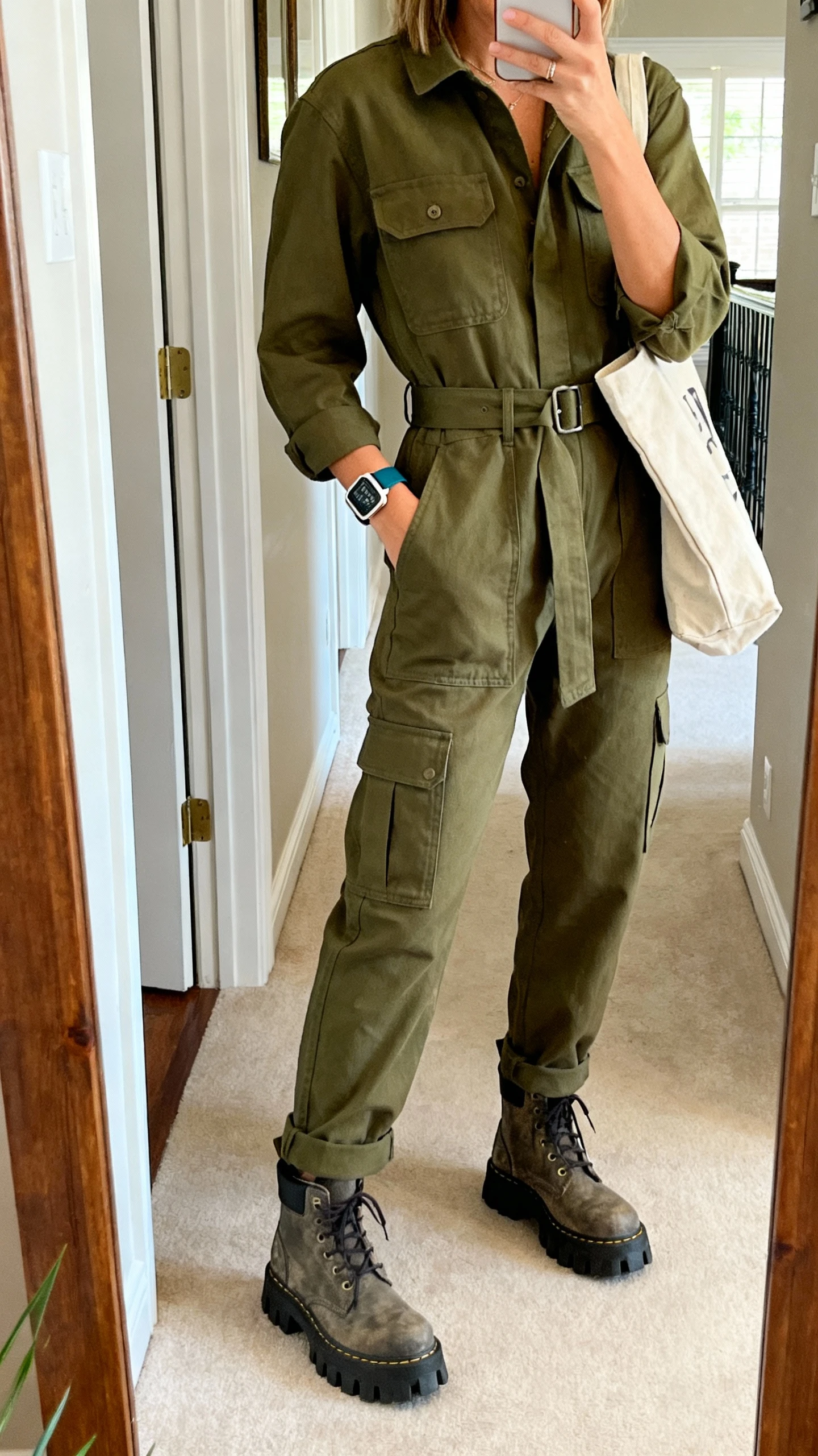 iPhone mirror selfie of a woman wearing an olive utility jumpsuit with cargo pockets and a belted waist, chunky lug-sole boots, canvas tote, sporty watch, face not visible, cropped at shoulders, entryway mirror, natural daylight, iPhone photo quality.