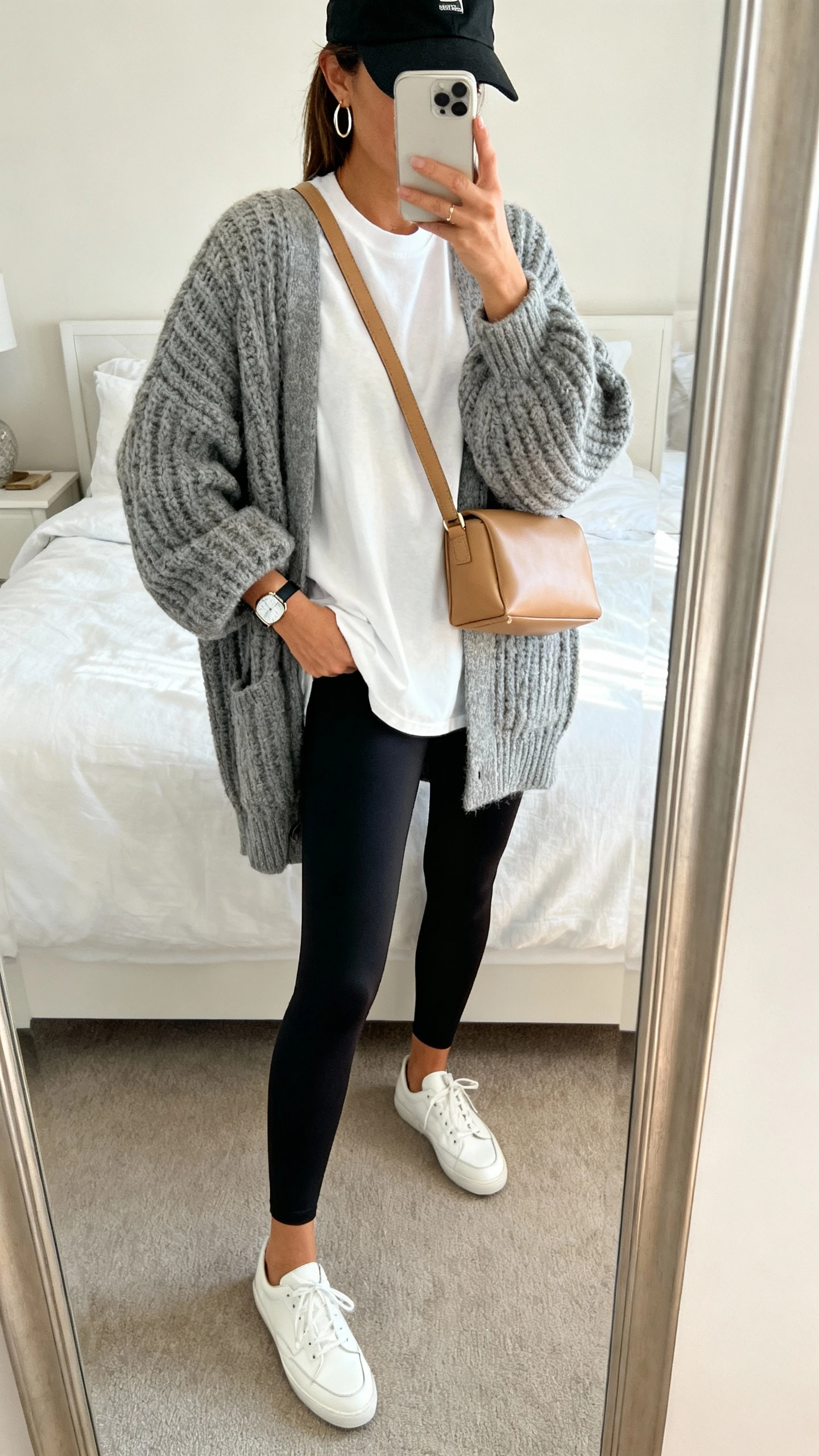 iPhone mirror selfie of a woman wearing an oversized chunky knit cardigan in heather gray, high-waisted matte black leggings, white low-top leather sneakers, a white boxy crewneck tee, a tan structured crossbody, simple hoop earrings, slim watch, and a black baseball cap, face not visible, cropped at neck, bedroom mirror with natural morning light, iPhone photo quality.