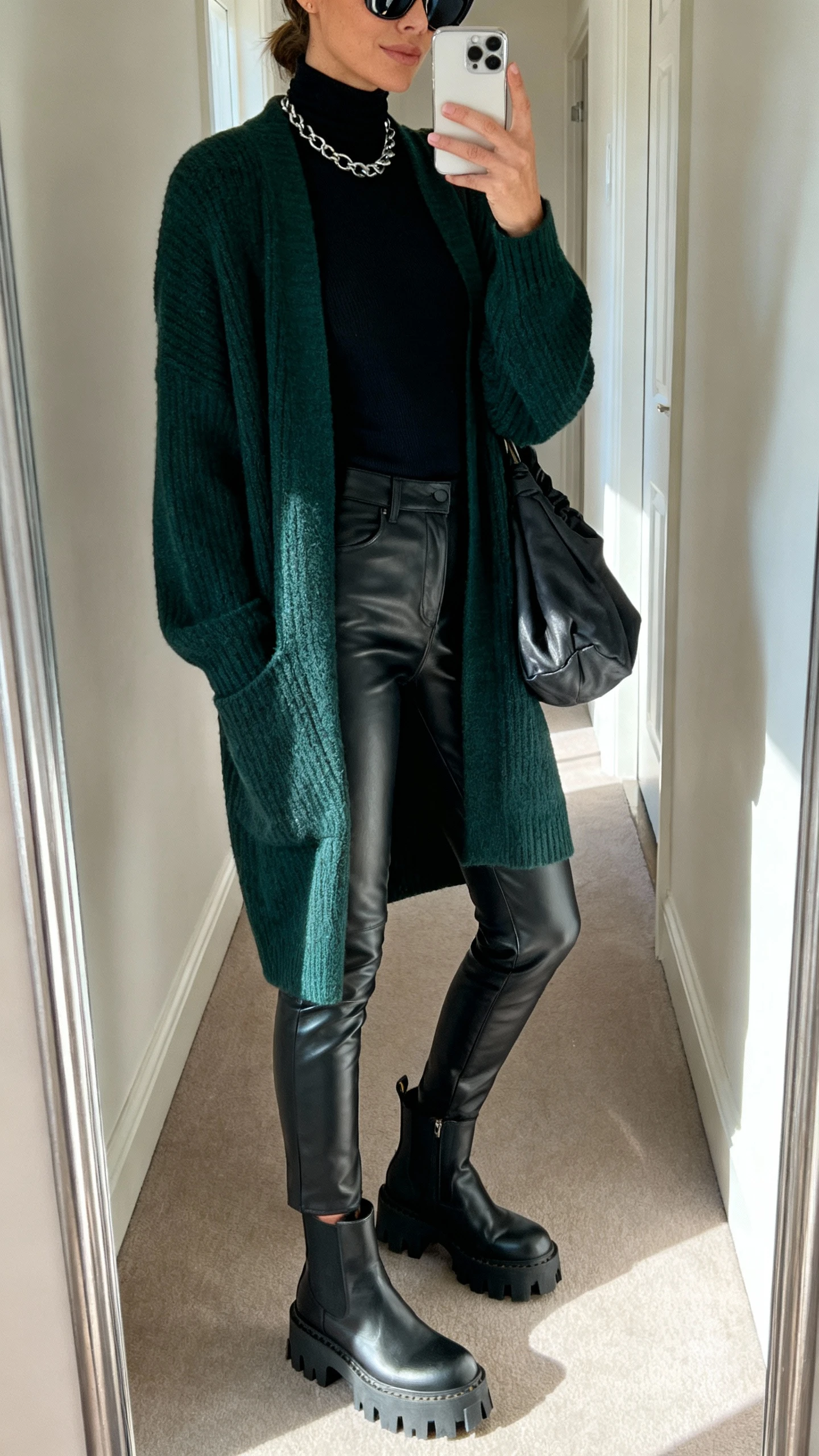 iPhone side-angle mirror selfie of a woman wearing a longline deep forest green cardigan, black faux leather slim pants, a fitted black turtleneck, chunky lug-sole ankle boots, a slouchy hobo bag, statement sunglasses, and a chain necklace, face not visible, hallway mirror with natural daylight, iPhone photo quality.