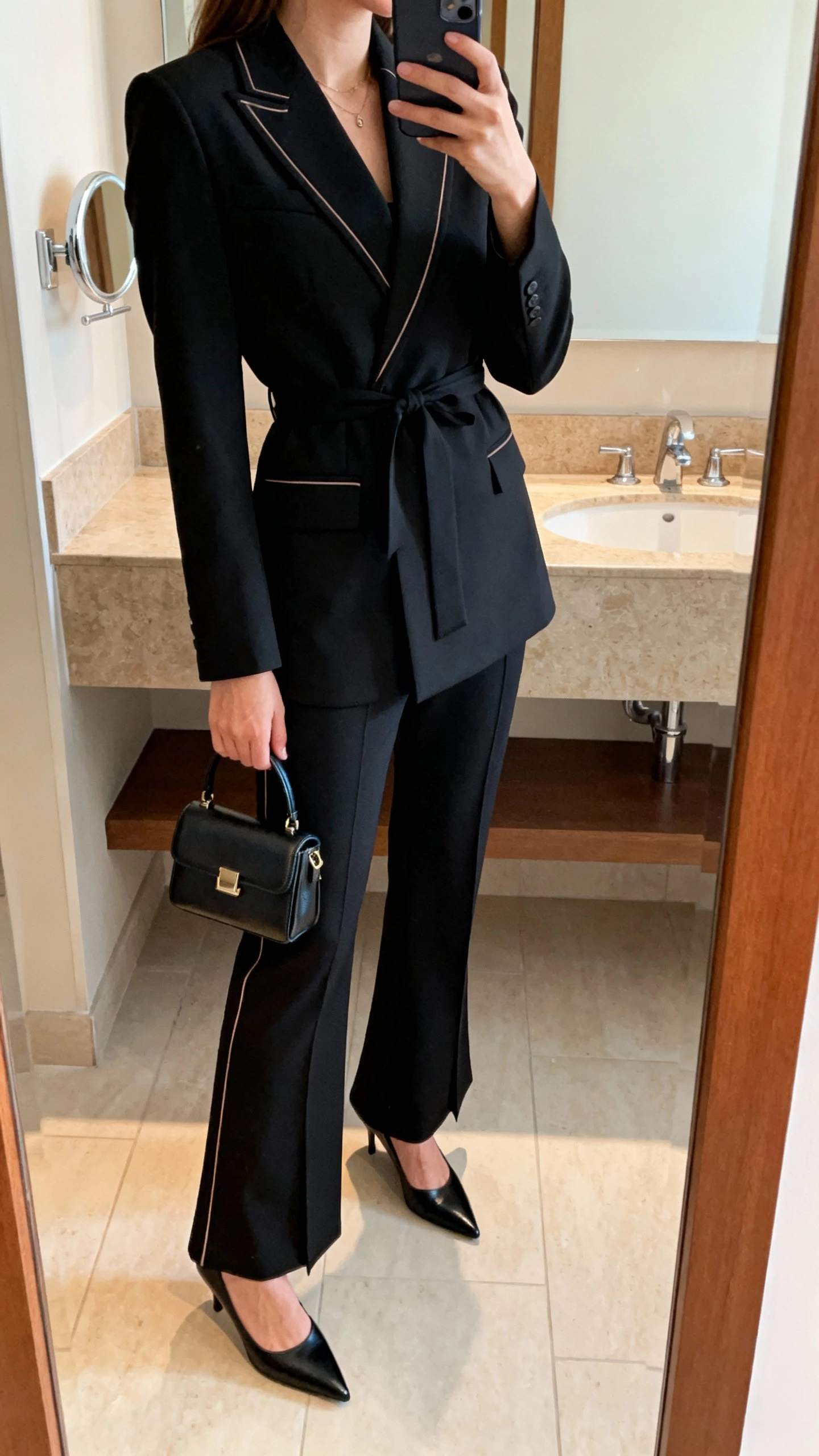 iPhone side-angle mirror selfie of a woman wearing a sharp black suit with a cinched waist, subtle tonal piping, and slightly flared trousers, black pointed pumps, and a structured mini bag, face not visible, bathroom mirror, soft warm lighting, iPhone photo quality.