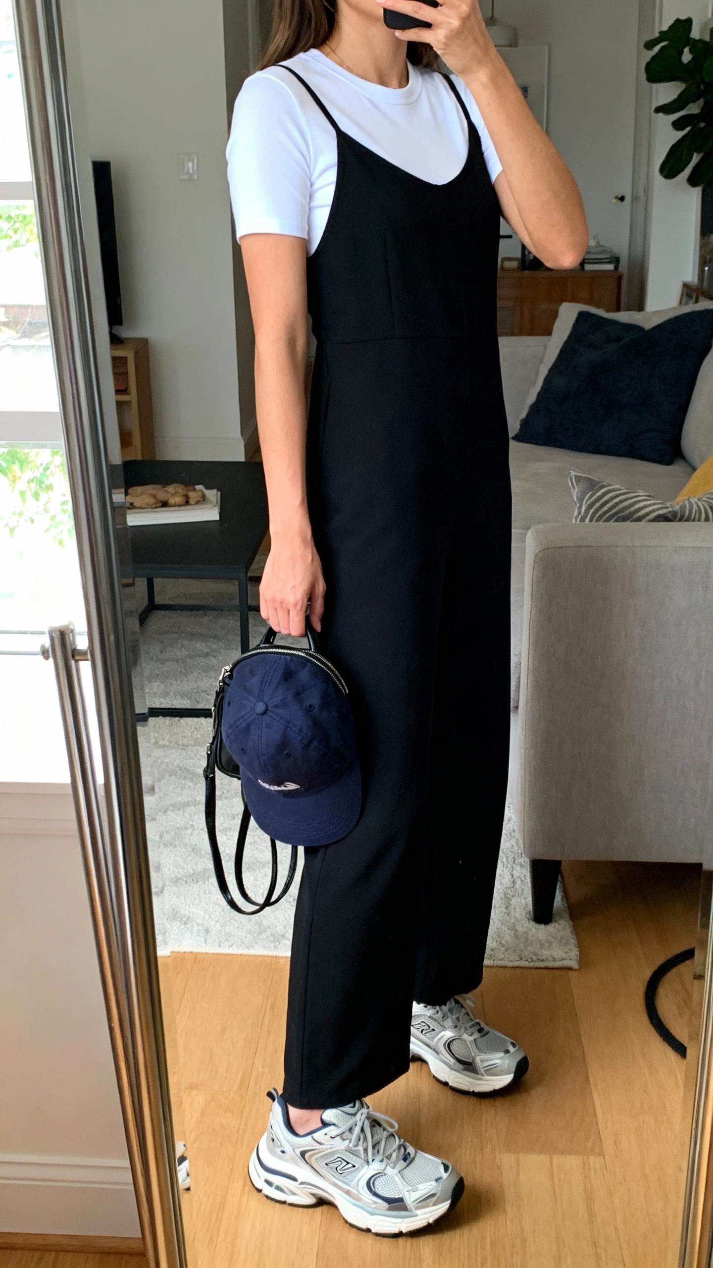 iPhone side-angle mirror selfie of a woman wearing a black tank jumpsuit layered over a white fitted crewneck T-shirt, chunky sneakers, baseball cap in hand, mini backpack, face not visible, cropped at neck, casual living room mirror, natural daylight, iPhone photo quality.