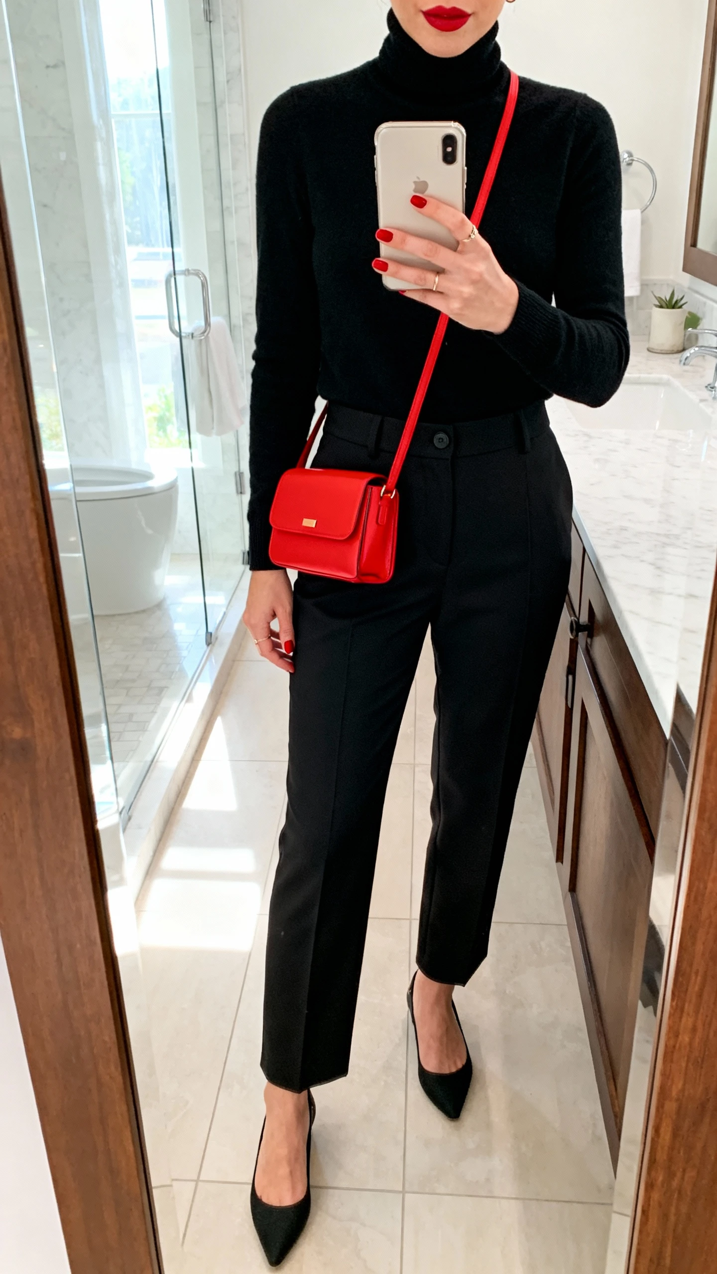 iPhone side mirror selfie of a woman wearing a monochrome set (black turtleneck and black tailored pants) with a bold red mini bag and matching lip-colored nails, minimalist pumps, face not visible, cropped at neck, bathroom mirror, bright indoor light, iPhone photo quality.