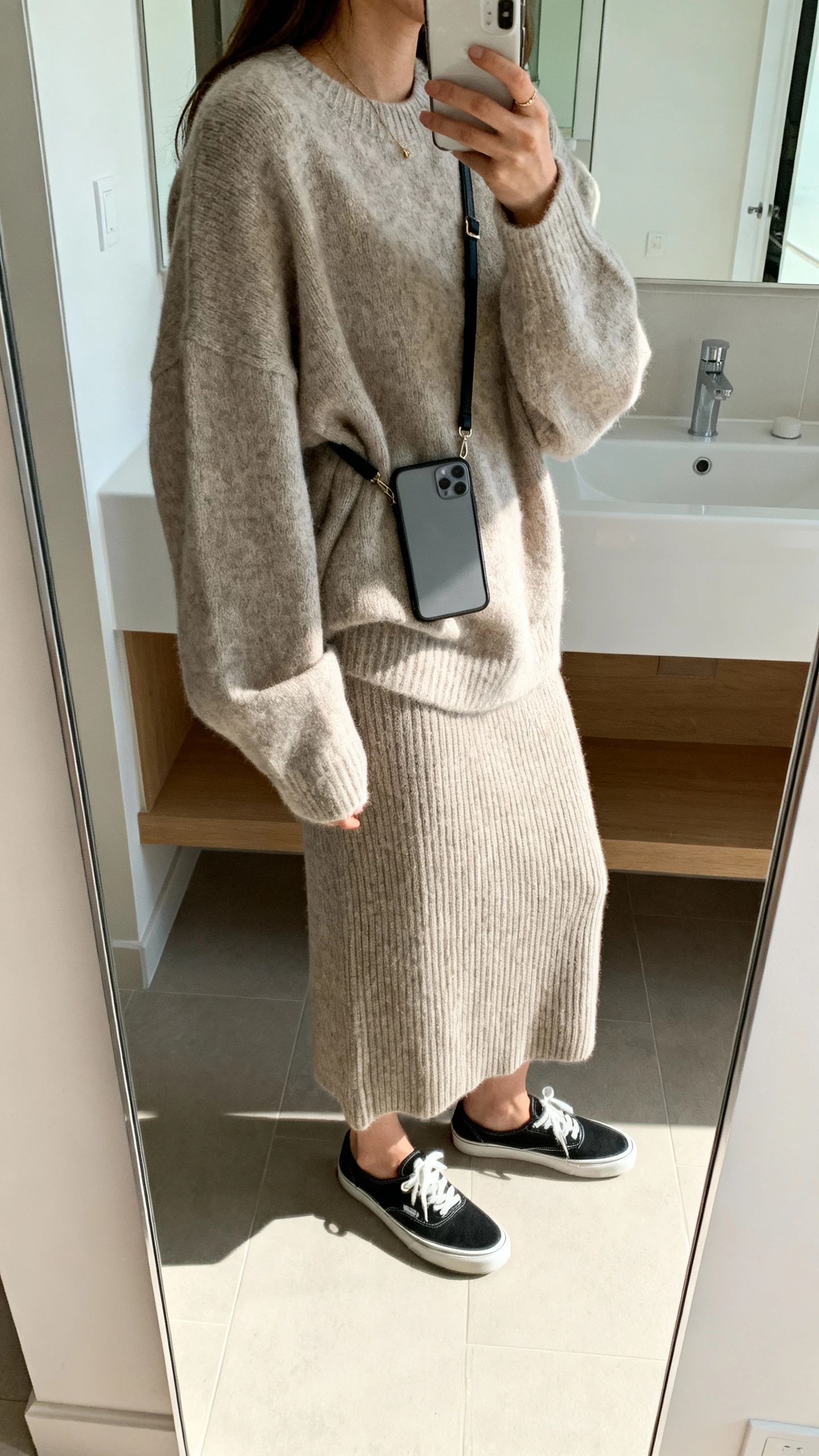 iPhone side-view mirror selfie of a woman wearing an oversized sweater with a knit midi skirt, slip-on sneakers, crossbody phone sling, face not visible, cropped at neck, bathroom mirror with bright indoor light, iPhone photo quality.