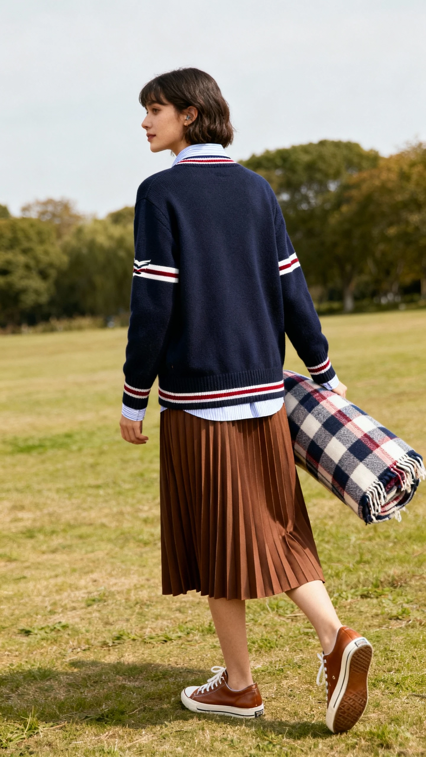 Park-side candid of a woman in a rowing-club cardigan layered over a collared shirt with a pleated midi skirt and leather sneakers, carrying a picnic blanket, face looking away, gentle daylight, iPhone photo quality.