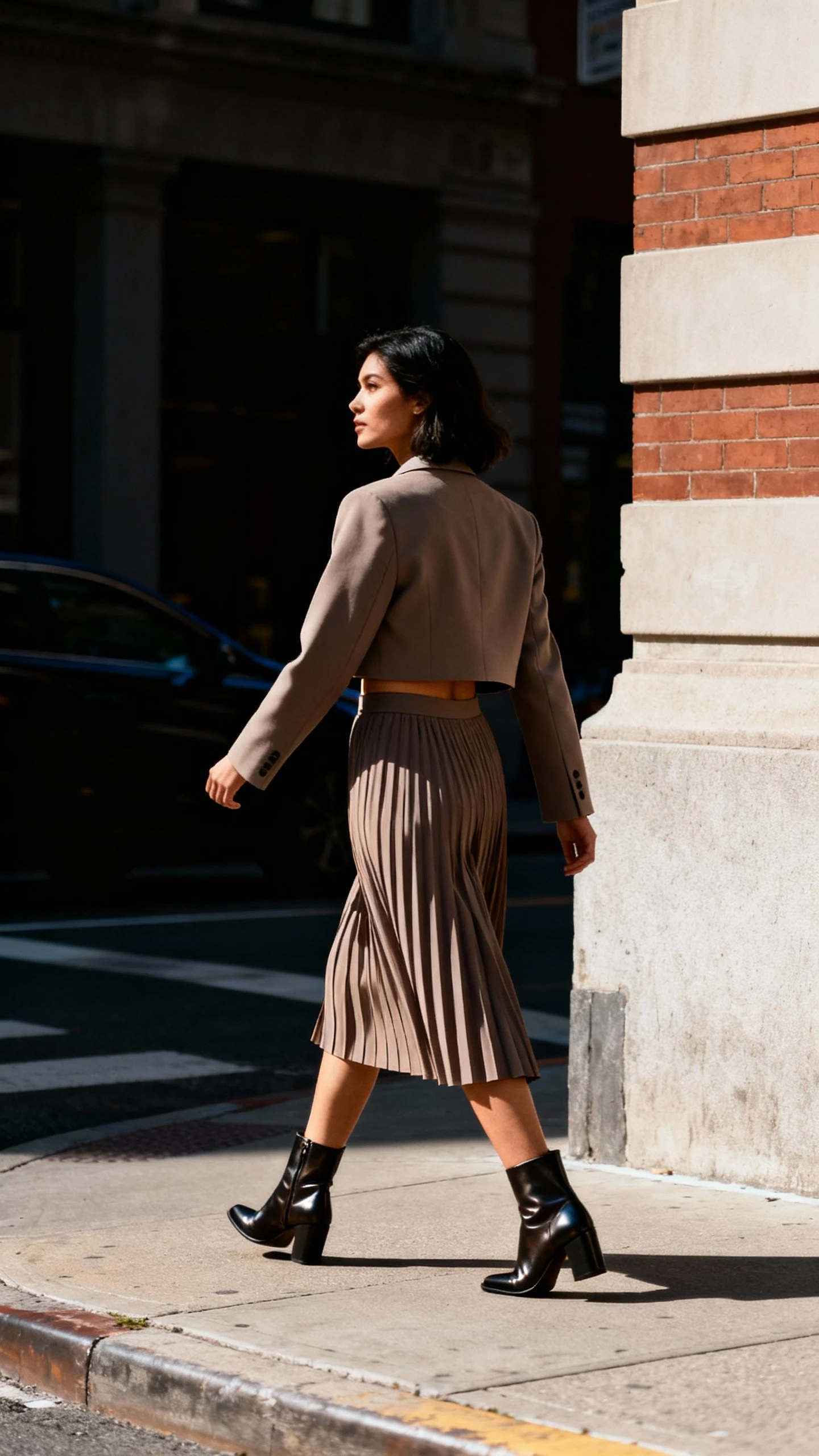 Street-style candid of a woman in a cropped blazer and high-waisted pleated skirt set, paired with sleek ankle boots, turning a corner on a city block, face in shadow looking away, afternoon daylight, iPhone photo quality, unstaged.