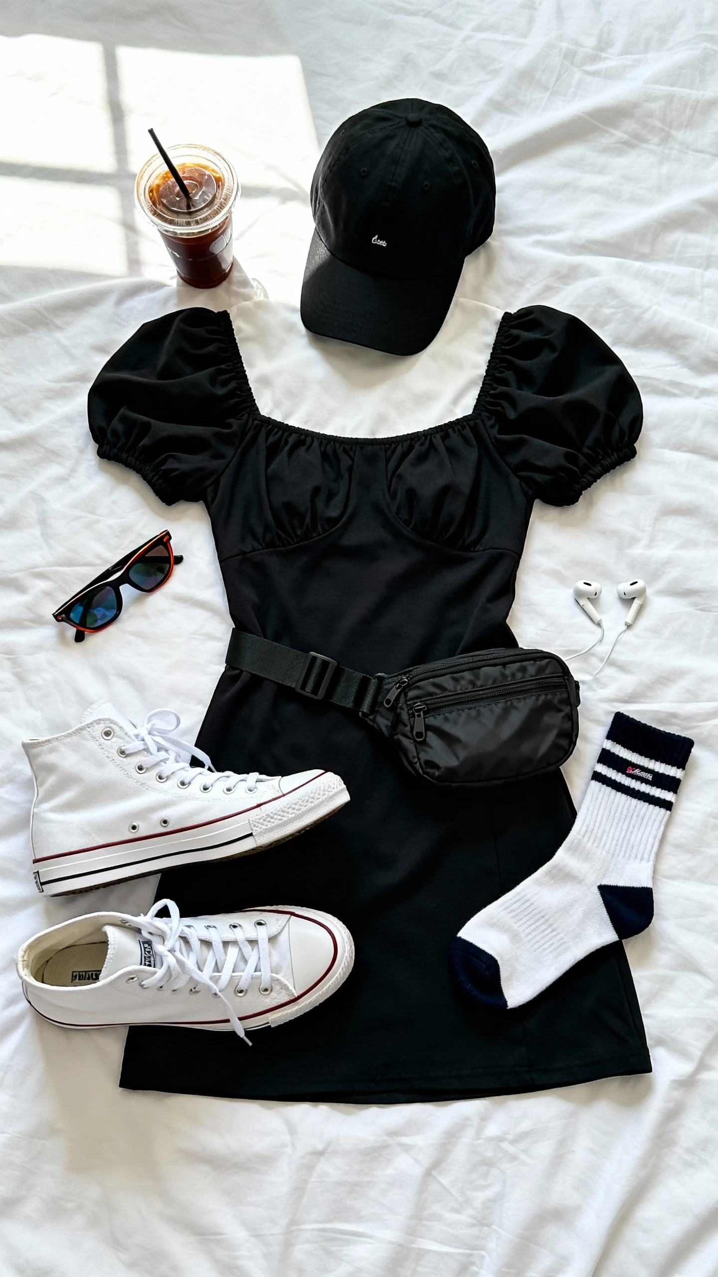 Top-down flat lay of a sporty-chic black puff sleeve dress with white high-top sneakers, black baseball cap, nylon belt bag, crew socks, and sporty sunglasses; a cold brew cup and wireless earbuds as props on a white bedspread, bright natural window light, no person visible, clean organized composition, iPhone photo aesthetic.